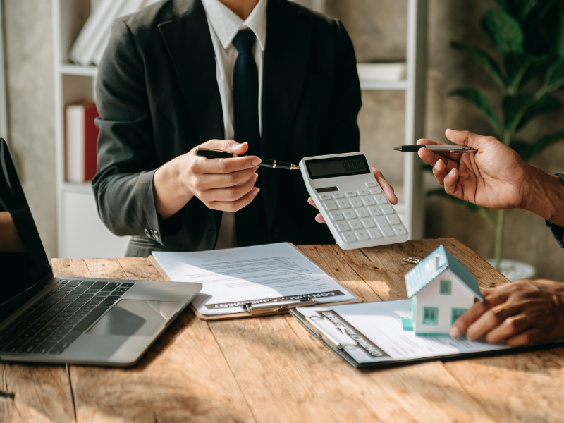 A mortgage broker shows the display of a calculator to a client, documents, a miniature house, and a laptop on the table
