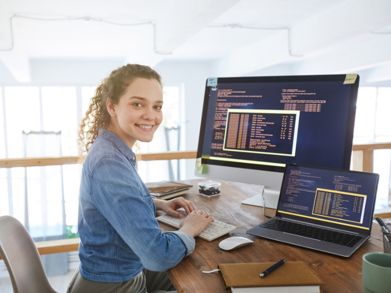 A female IT professional smiles at the camera, her monitor and laptop both showing coding programs