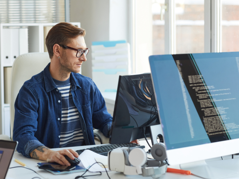 An IT professional in glasses works on his computer, another monitor showing lines of code