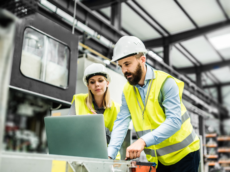 Two employees at a manufacturing plant wearing safety gear look at a laptop