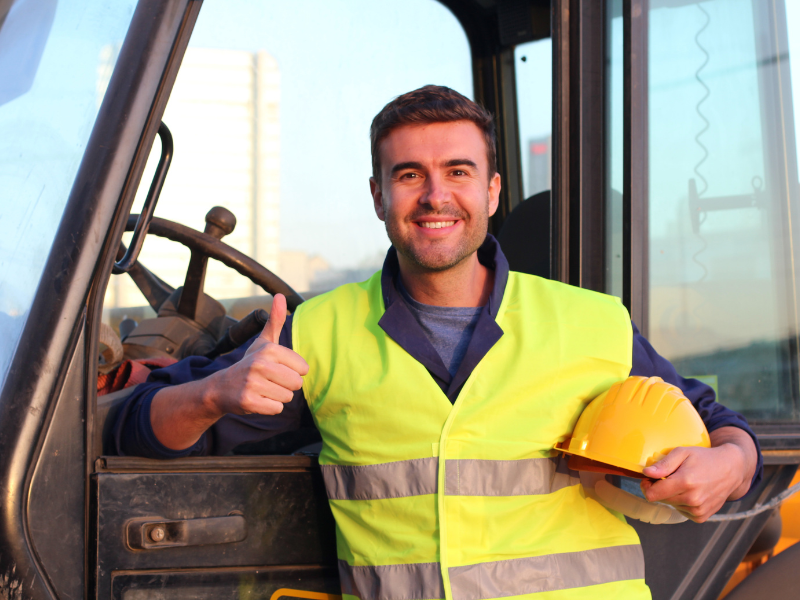 A crane driver leans on the door of the crane car, posing with his thumbs up and his other hand holding his yellow hard hat