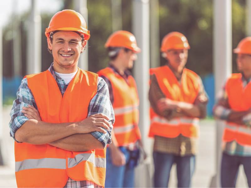 A construction worker in orange safety gear poses with his arms crossed and smiling at the camera, three more construction workers in the background