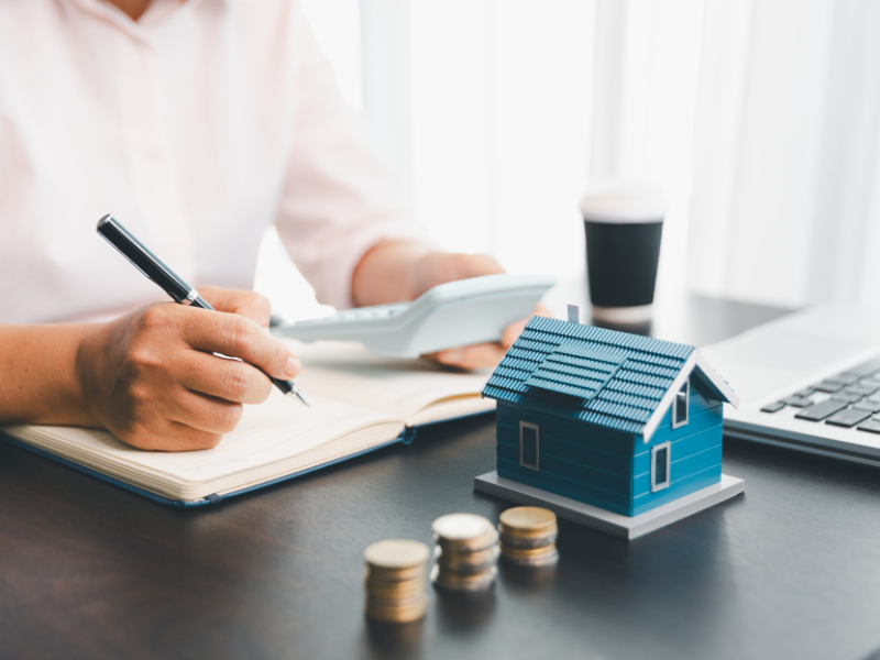 Cropped photo of a man writing calculations on a notebook, a miniature house and piles of coins in front of him, a man getting a loan secured against property