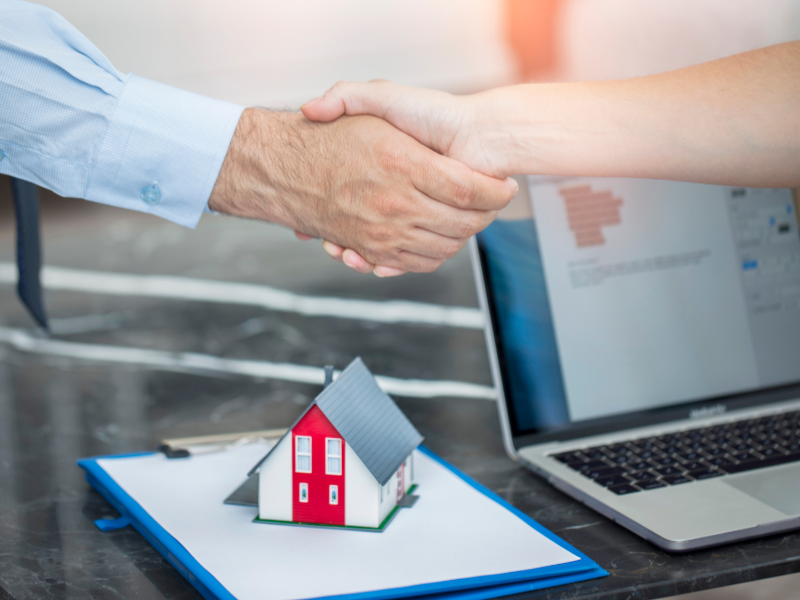 Two hands shaking, a clipboard with a document, a laptop, and a miniature model house below