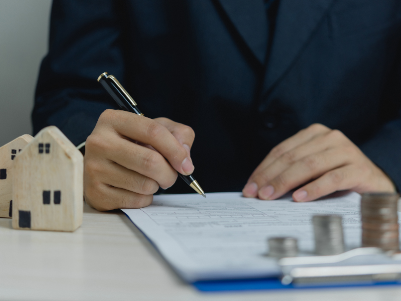 A man filling out a loan application form, mini wooden houses and piles of coins next to him