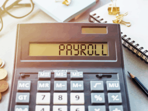 A black calculator displaying the word “payroll”, a notebook, pencil, pile of coins, glasses, and sticky notes placed around the calculator