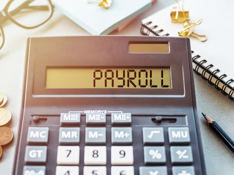 A black calculator displaying the word “payroll”, a notebook, pencil, pile of coins, glasses, and sticky notes placed around the calculator