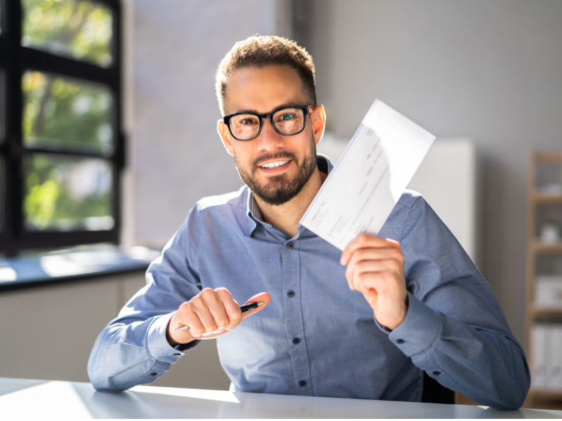 A man in glasses smiles as he holds a paycheck, employer about to hand a contractor his paycheck