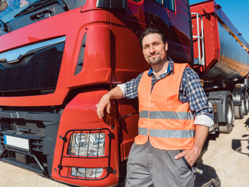 A transport truck driver wearing an orange high visibility vest poses next to his red freight truck