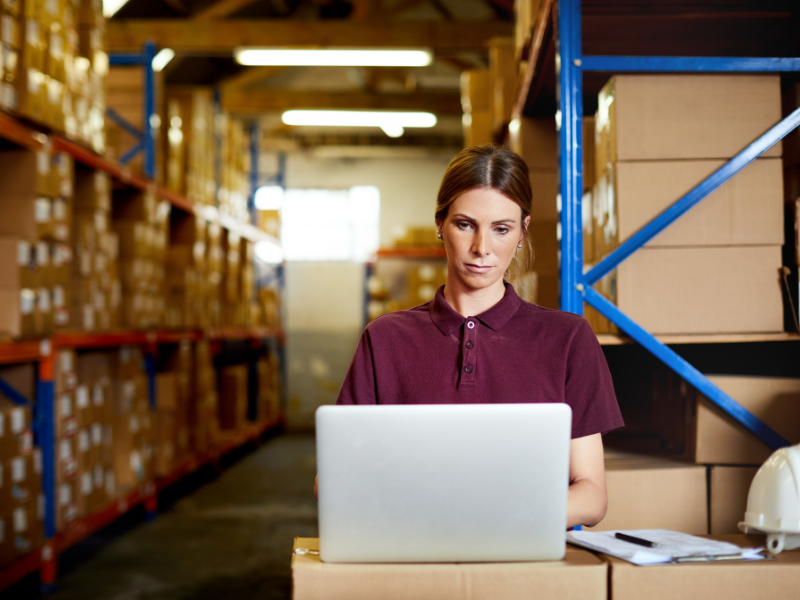 A woman uses her laptop inside a warehouse full of boxes, a supplier tracking payments from clients