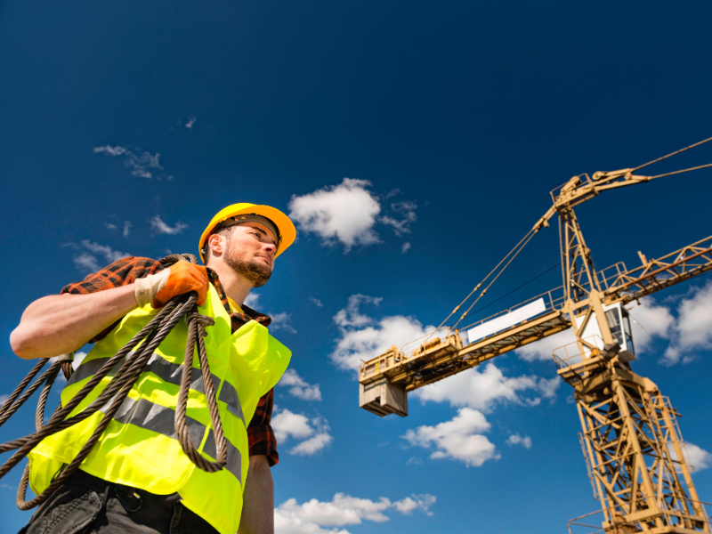 A construction worker holding a rope, a large crane and clear blue skies in the background