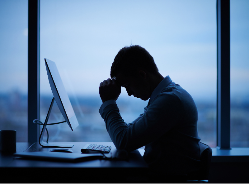 Silhouette from the light of the window, a man in front of his computer with his head hanging low and hand supporting his forehead, business owner stressed with high tax debt