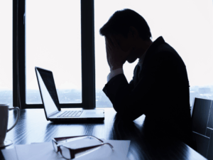 A silhouette of a man hanging his head low with his hands over his face, against the light of an office window, a man stressed about tax debt