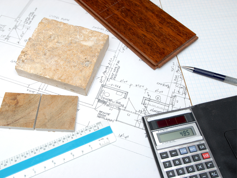Renovation plans, blueprints, wood and tile samples, a ruler, pen, and calculator on a table, calculating fitout costs