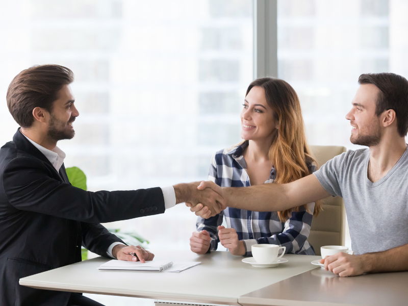 A man seated next to a woman shakes hands with another man in a suit, business owners agreeing on a loan with a lender or loan broker