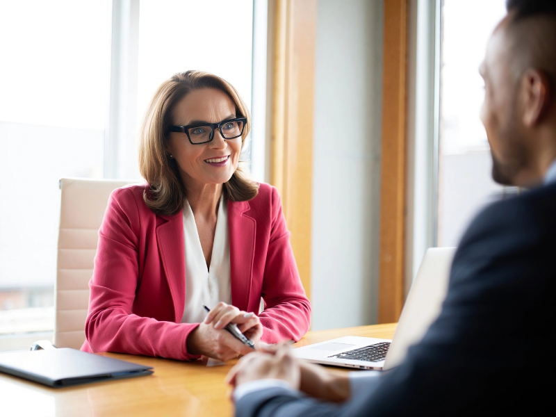 A woman in glasses cheerfully speaks with a man in a suit, a mortgage broker discusses refinancing with a business owner