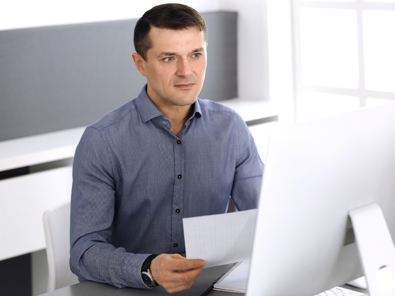 A man in a white and sleek office holds a document while reading the display of a computer monitor, business owner considering funding options for emergencies