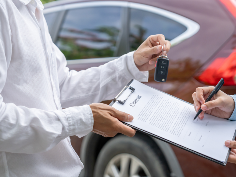 Cropped photo of a dealer holding a contract and car keys while a business owner signs the contract for a car sale