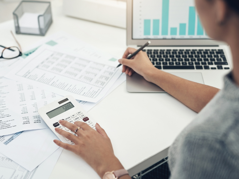 Cropped photo of a woman calculating payroll for employees, woman writing on documents while using a calculator, an open laptop beside her