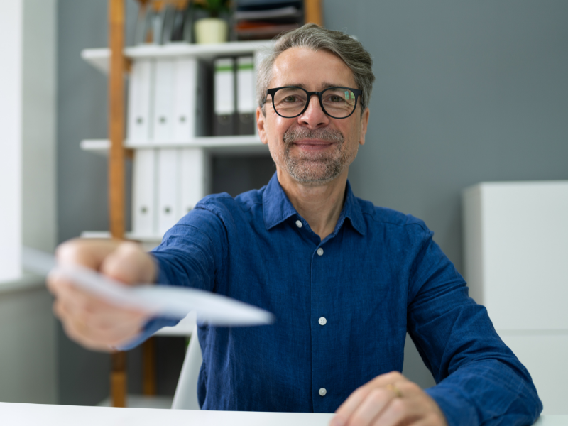 An employer happily hands over an employee’s paycheck, a man in glasses handing a paycheck towards the camera