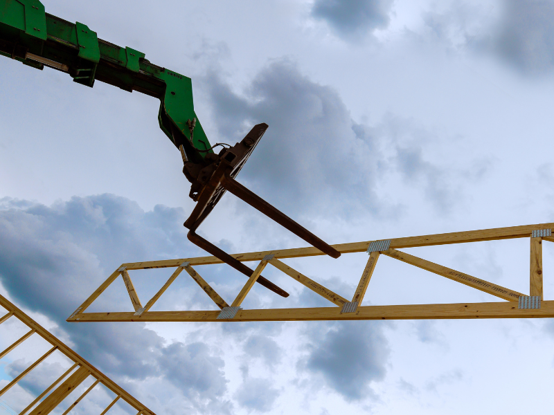 Heavy telehandler machinery lifts wooden truss above construction site under cloudy sky at dusk