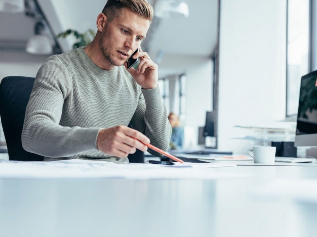 A man in an office talking on the phone while reading a document on his desk, business owner speaking with accountant
