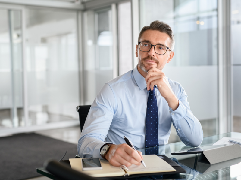 A man in glasses seated at his office table jots down notes, a business owner thinking of applying for an unsecured business loan